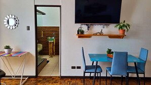A modern room with a TV mounted on the wall, a dining table with chairs, a small side table with a potted plant, and a mirror on the wall. - 714 Studios (San José)