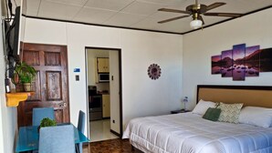 A bedroom with a bed, a desk, and chairs. There is a ceiling fan and a wall with a painting. - 714 Studios (San José)