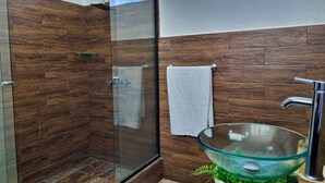 A modern bathroom with a glass shower enclosure, a glass sink with greenery, and a wooden wall. - 714 Studios (San José)