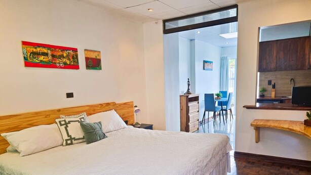 A hotel room with a large bed, a wooden headboard, a desk with a chair, and a kitchenette area. - 714 Studios (San José)