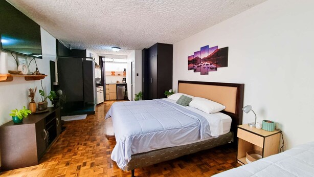 Une chambre avec un plancher en bois, un lit avec une literie blanche, une table de chevet avec une lampe et une œuvre d’art accrochée au mur.