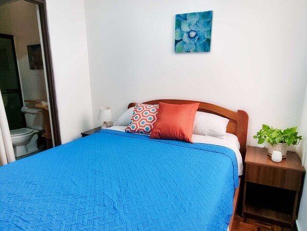 A bedroom with a bed, a bedside table, a nightstand, a potted plant, and a bathroom visible through an open door. - 714 Studios (San José)