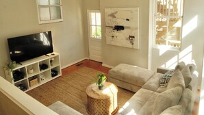 A living room with a flat-screen TV, a white sofa, a round wooden side table, and a large window with blinds.