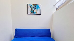 Una habitación pequeña y estrecha con un solo colchón azul sobre el suelo, una pared blanca y un cuadro abstracto enmarcado sobre el colchón.