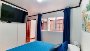 Un dormitorio con cama, televisor montado en la pared, ventana con cortinas y puerta que da a otra habitación.
