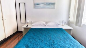 Une chambre à coucher comprenant un lit, deux tables de chevet, une commode, un tableau représentant un paysage marin et une fenêtre avec des rideaux.