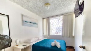 Une chambre à coucher avec un lit, une table de chevet, un ventilateur, une fenêtre avec des rideaux et un tableau au mur.