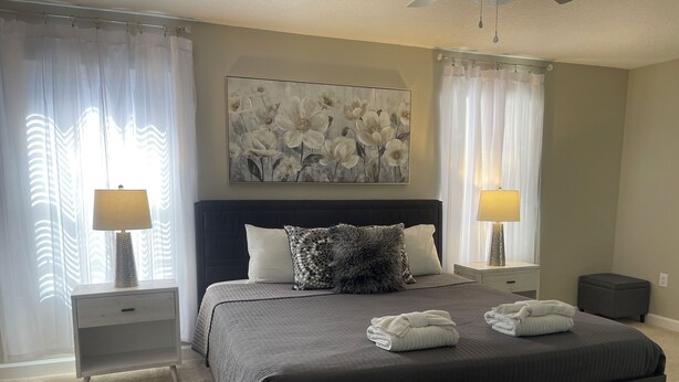 A bedroom with a large bed, two bedside tables, a headboard, a painting, and a window with curtains. - Magical Kingdom of Champions, 6-bed pool home golfing resort close to Disney! (Davenport)