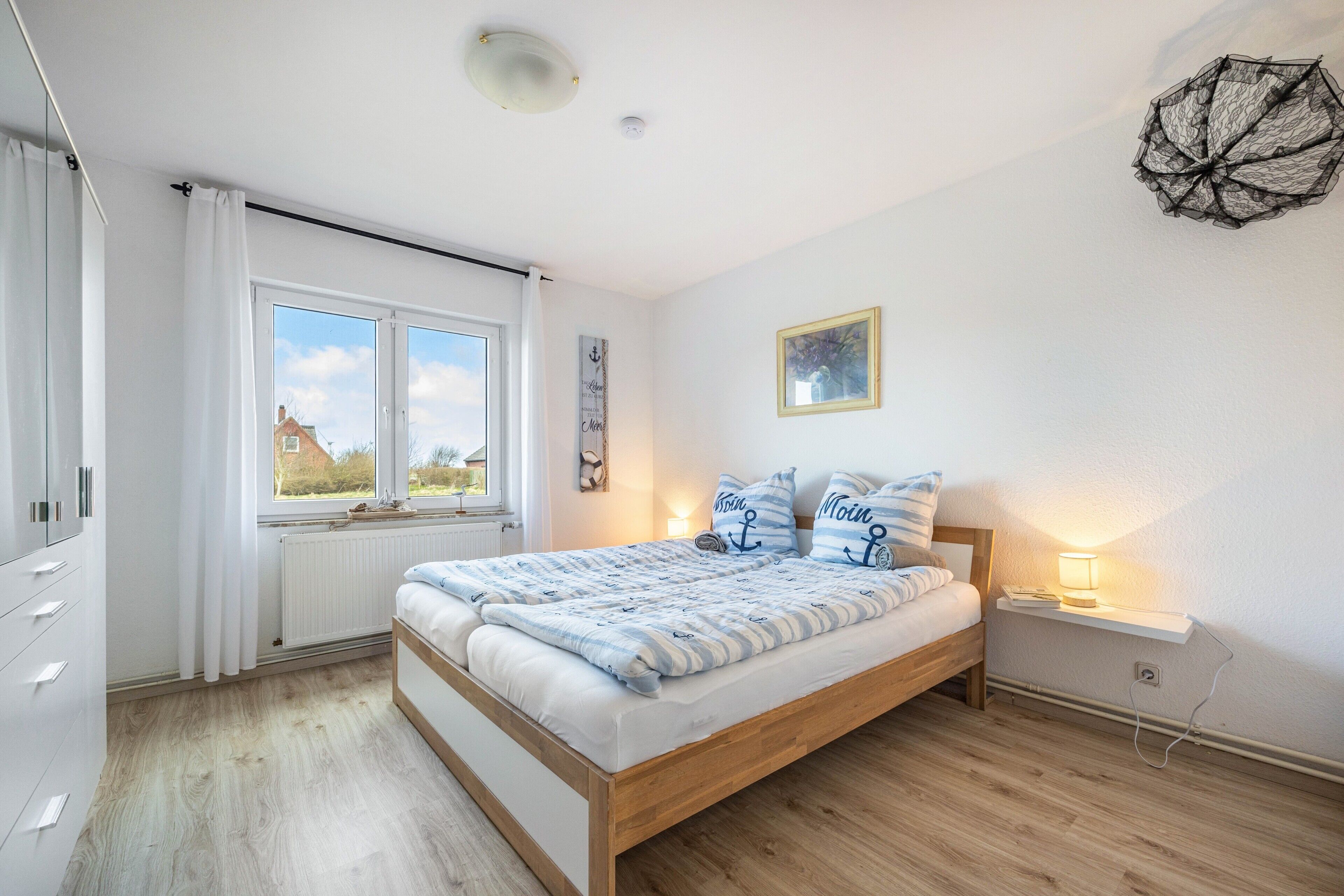 A bedroom with a wooden bed, white curtains, a window with a view, a bedside lamp, and a wall-mounted light fixture.