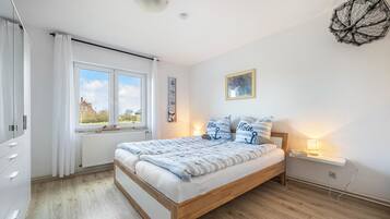 A bedroom with a wooden bed, white curtains, a window with a view, a bedside lamp, and a wall-mounted light fixture.