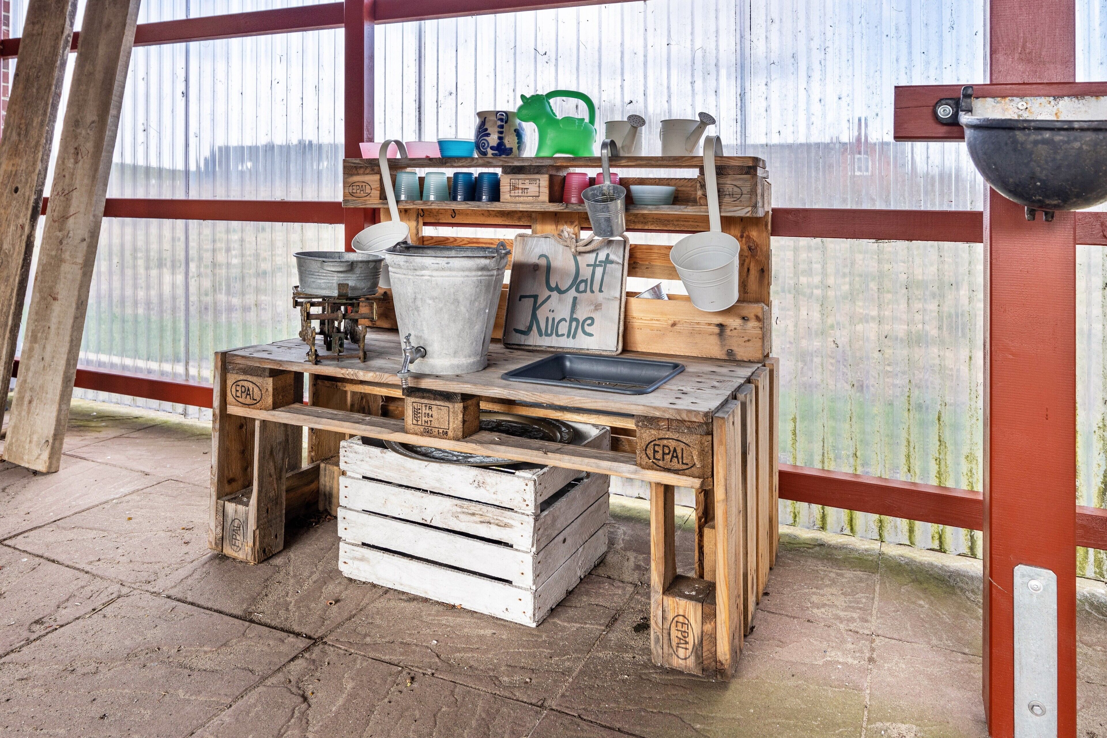 A rustic outdoor kitchen setup with wooden crates, metal pots, and a sign reading 'Watt Küche.'