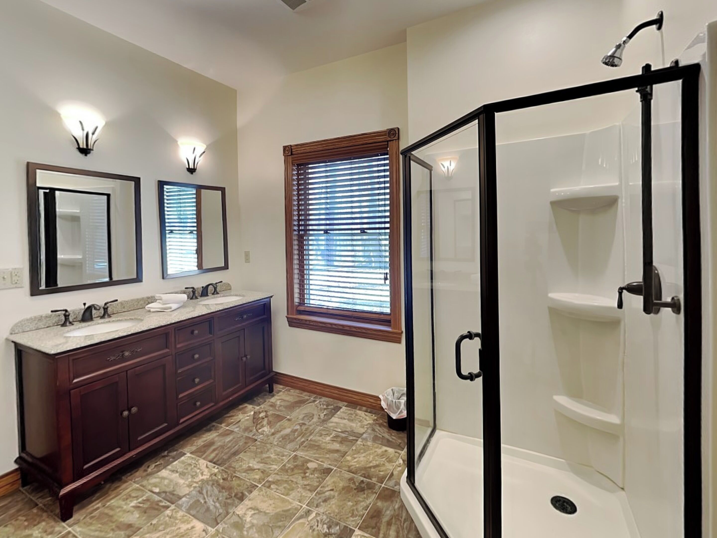 A bathroom with a double vanity, two wall-mounted mirrors, a window with blinds, and a walk-in shower with a black frame.