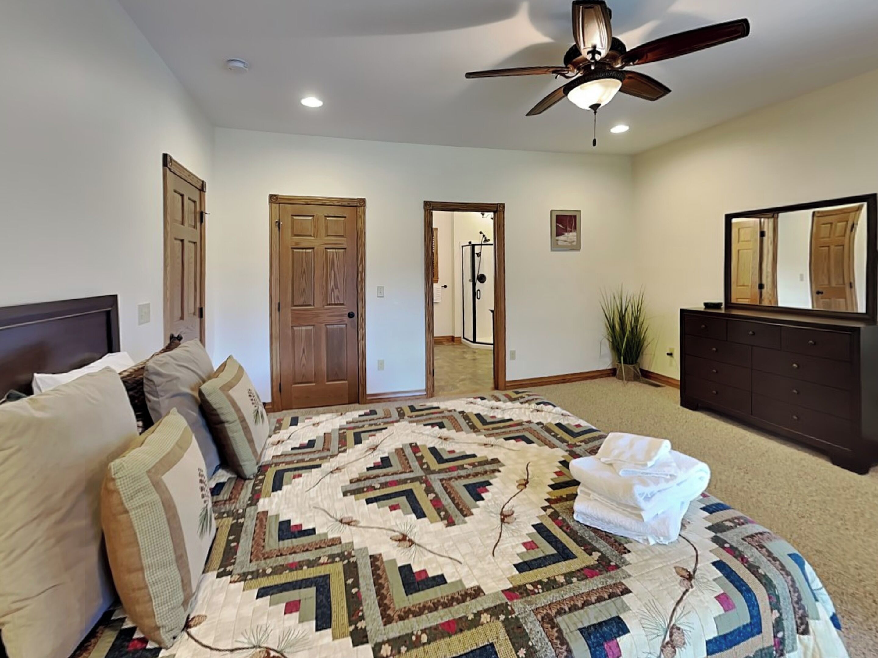 A bedroom with a large bed, a ceiling fan, a dresser, and a door leading to another room.