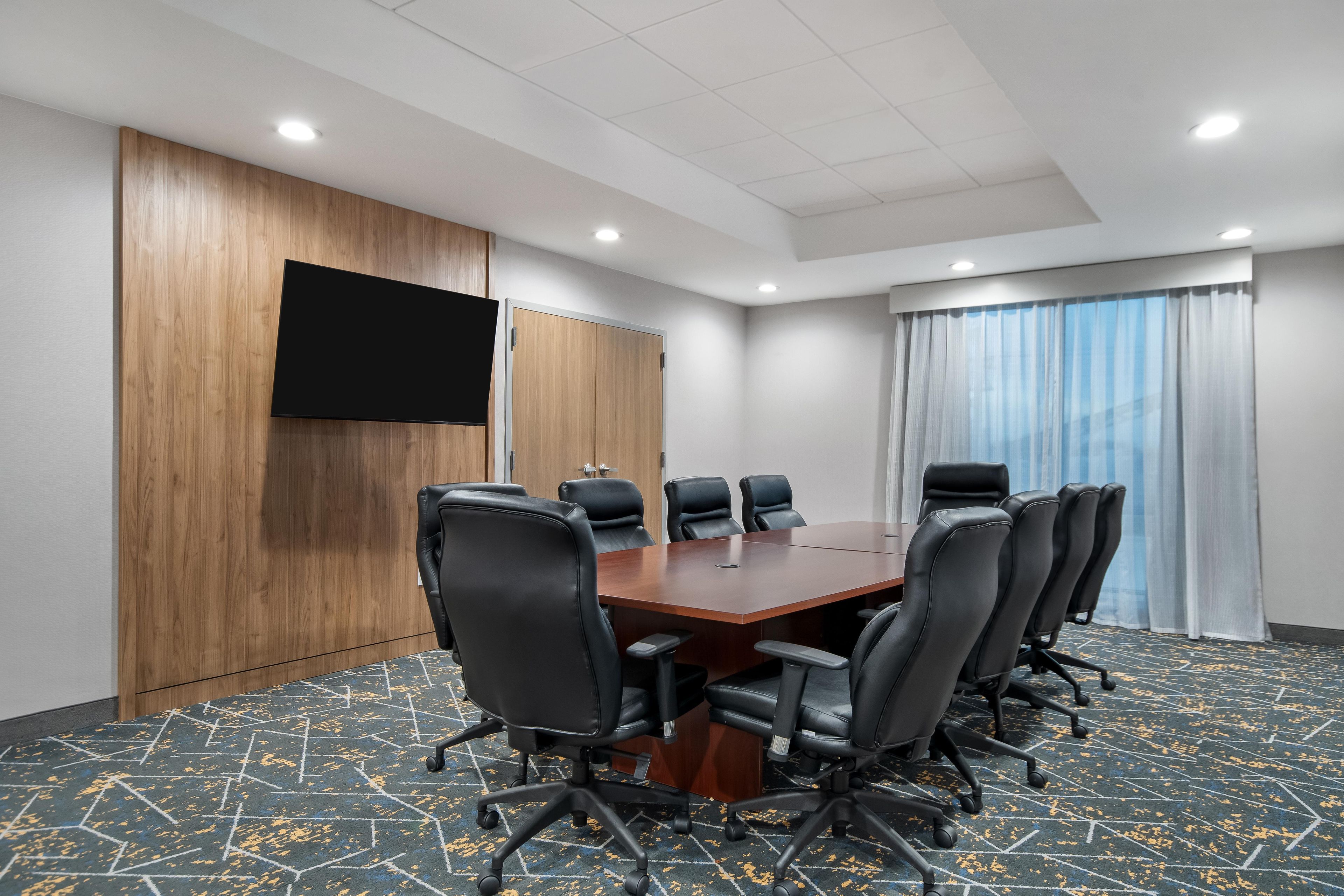 A conference room with a wooden panel wall, a flat-screen TV, a large conference table, and black leather chairs.