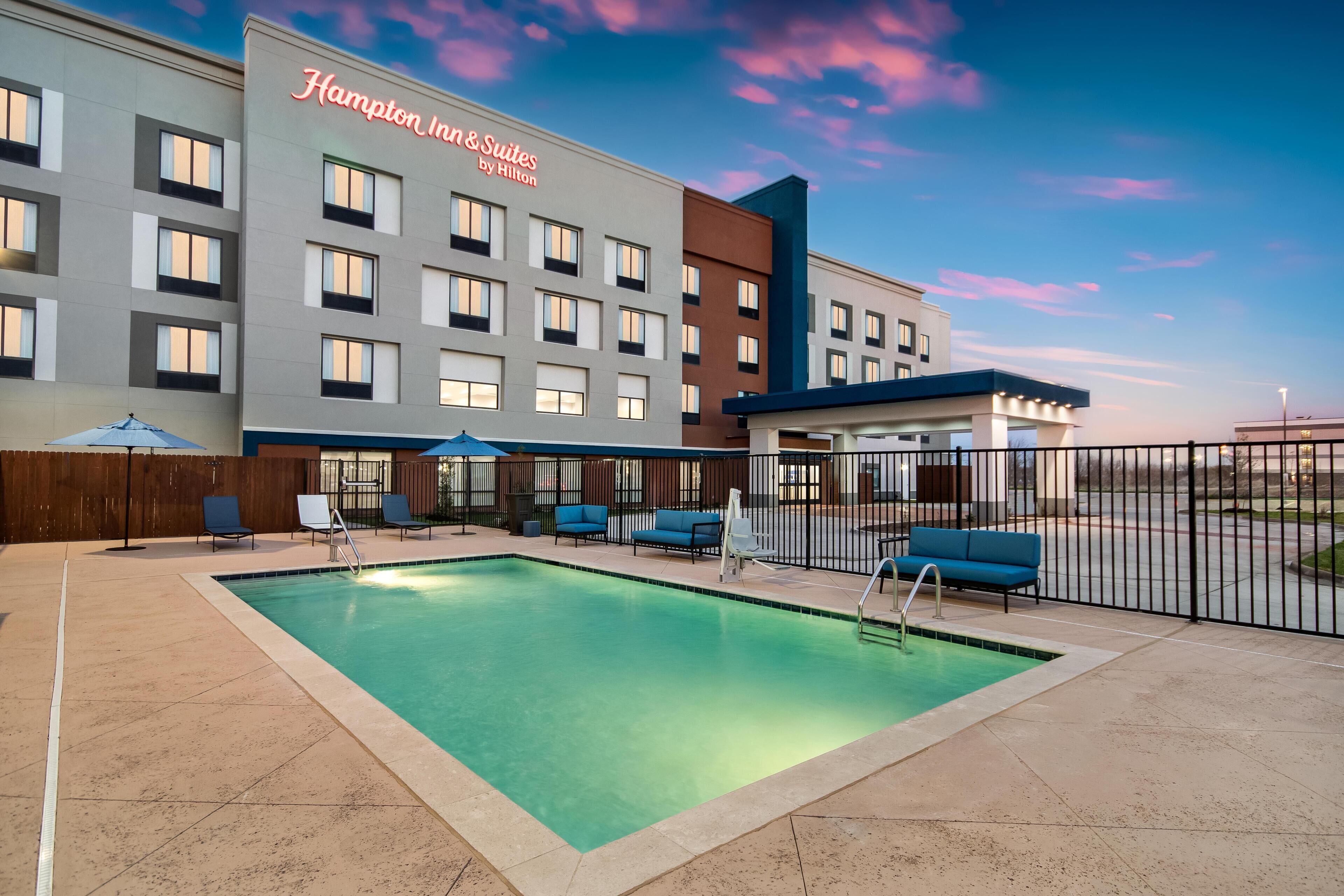 A hotel with a swimming pool, outdoor seating, and a clear sky.