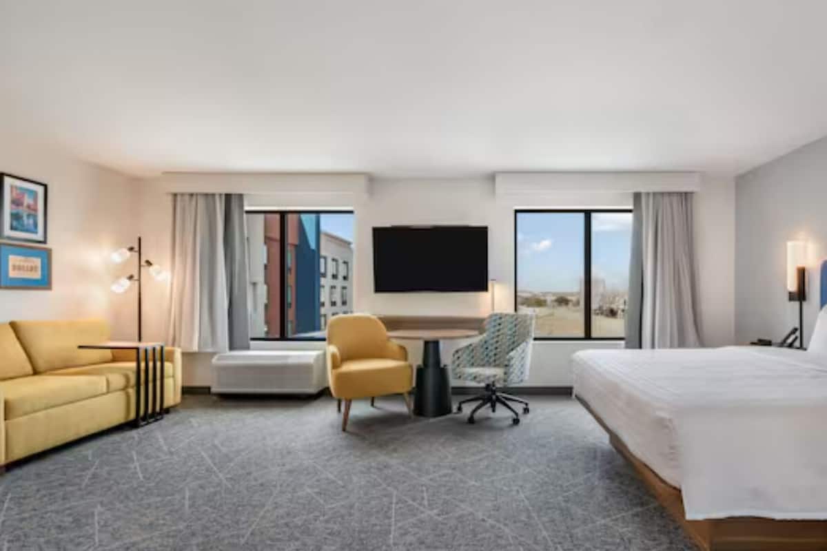 A hotel room with a bed, a sofa, a desk, a chair, a TV, and large windows.