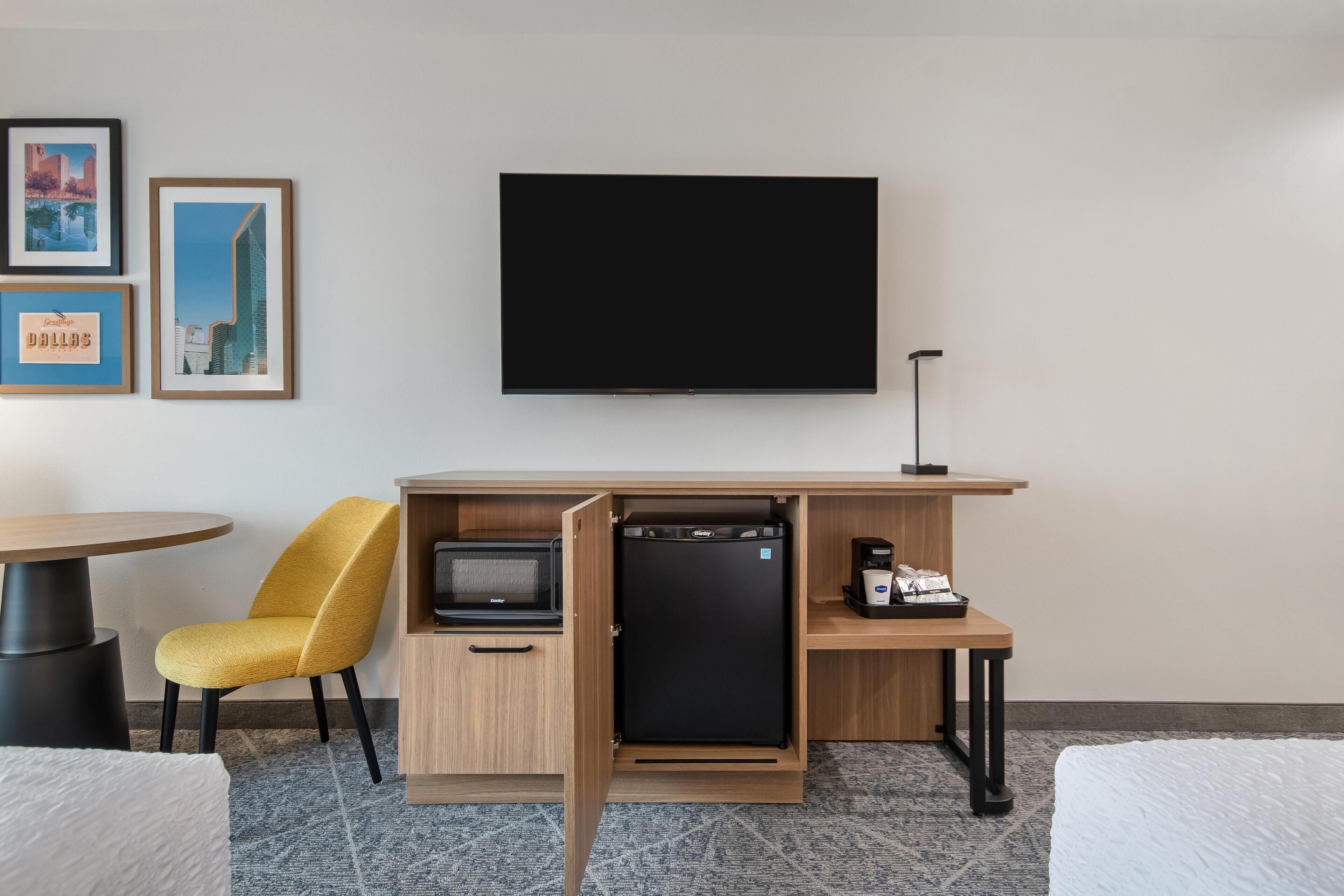 A compact hotel room with a bed, a desk with a flat-screen TV, a small refrigerator, a microwave, a coffee maker, and a chair.