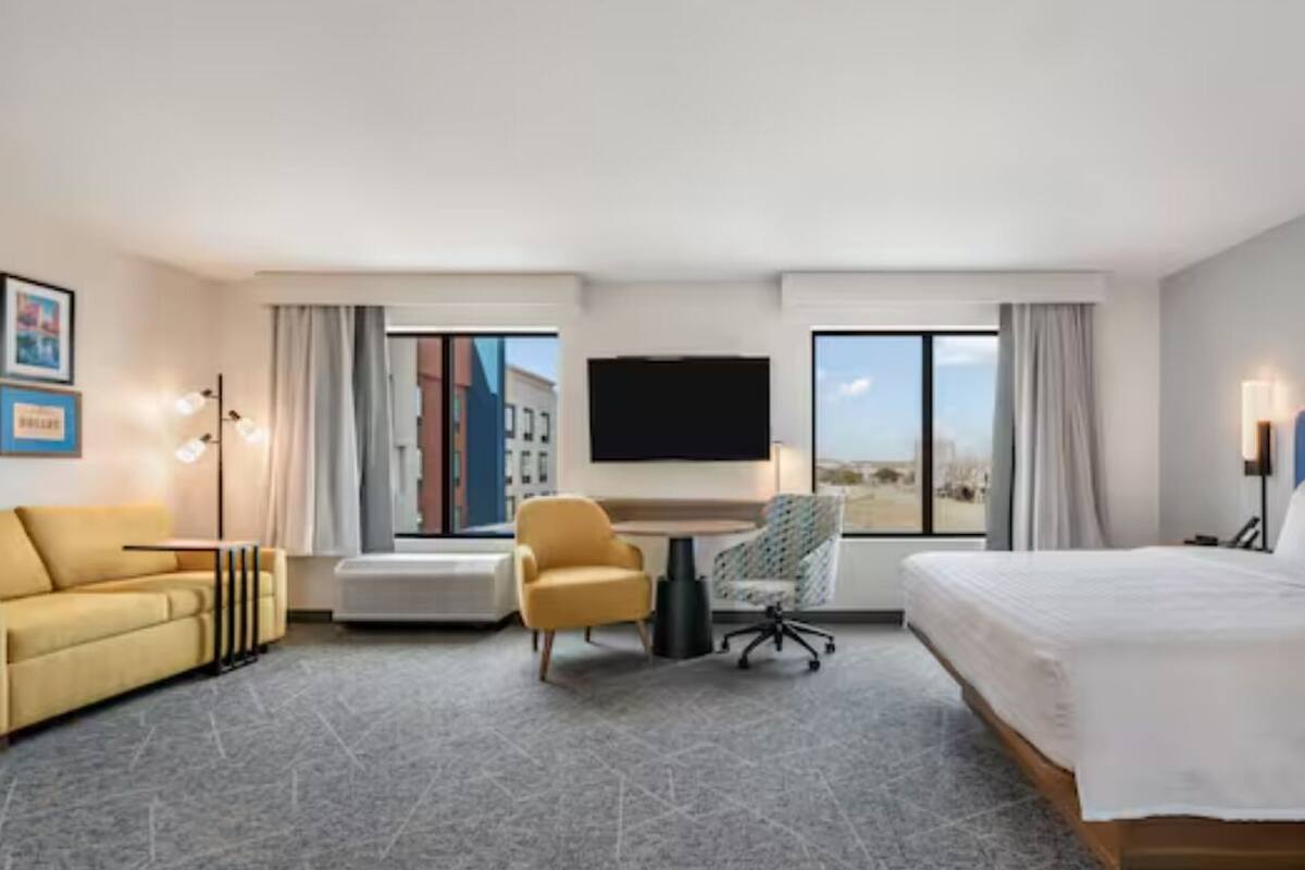 A hotel room with a bed, a sofa, a desk, a chair, a TV, and large windows.