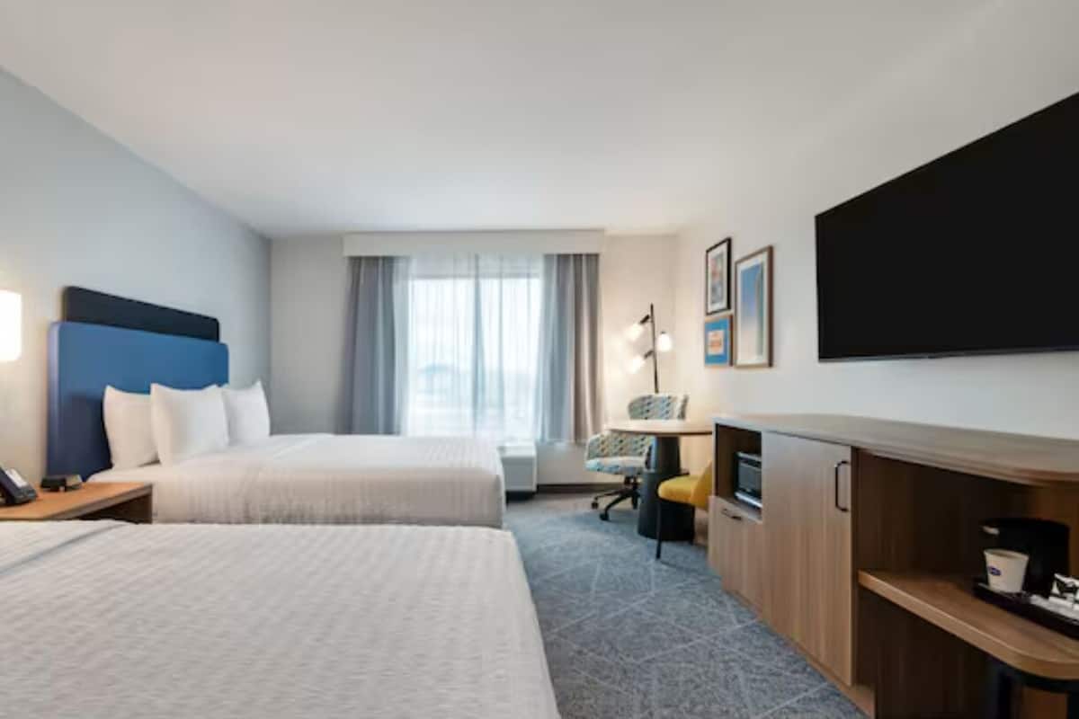 A hotel room with two beds, a desk, a flat-screen TV, and a window with curtains.