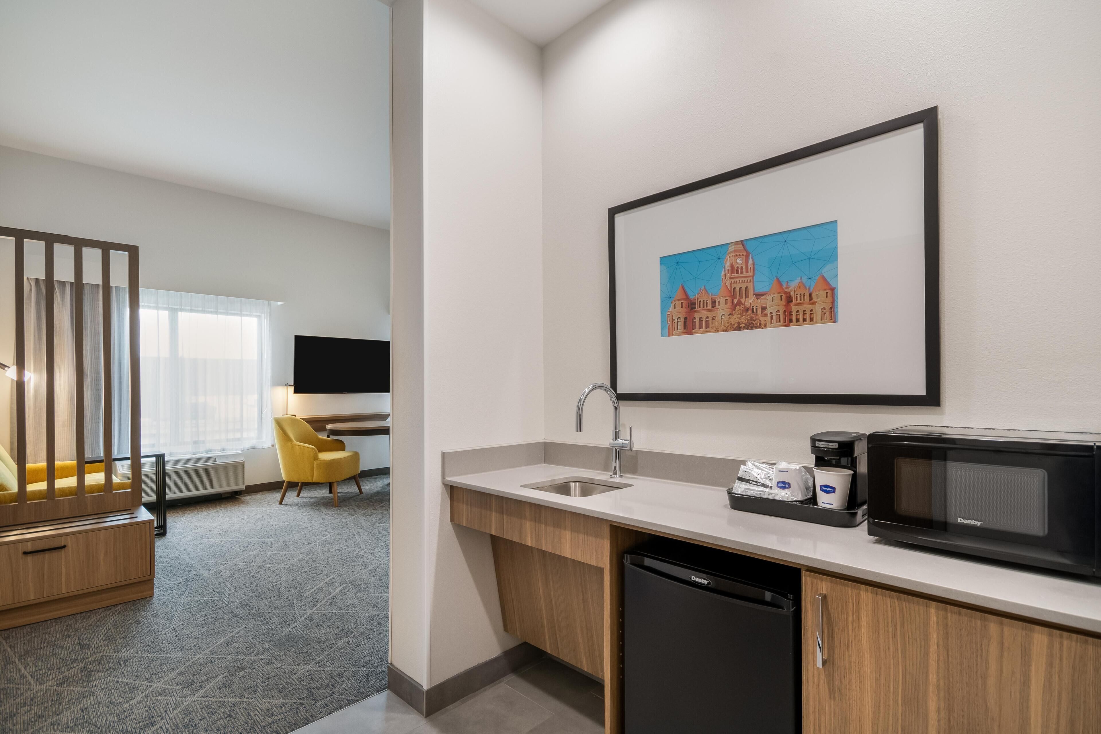 Une chambre d’hôtel moderne dotée d’une kitchenette, d’un téléviseur à écran plat, d’un fauteuil jaune et d’un tableau représentant un bâtiment.