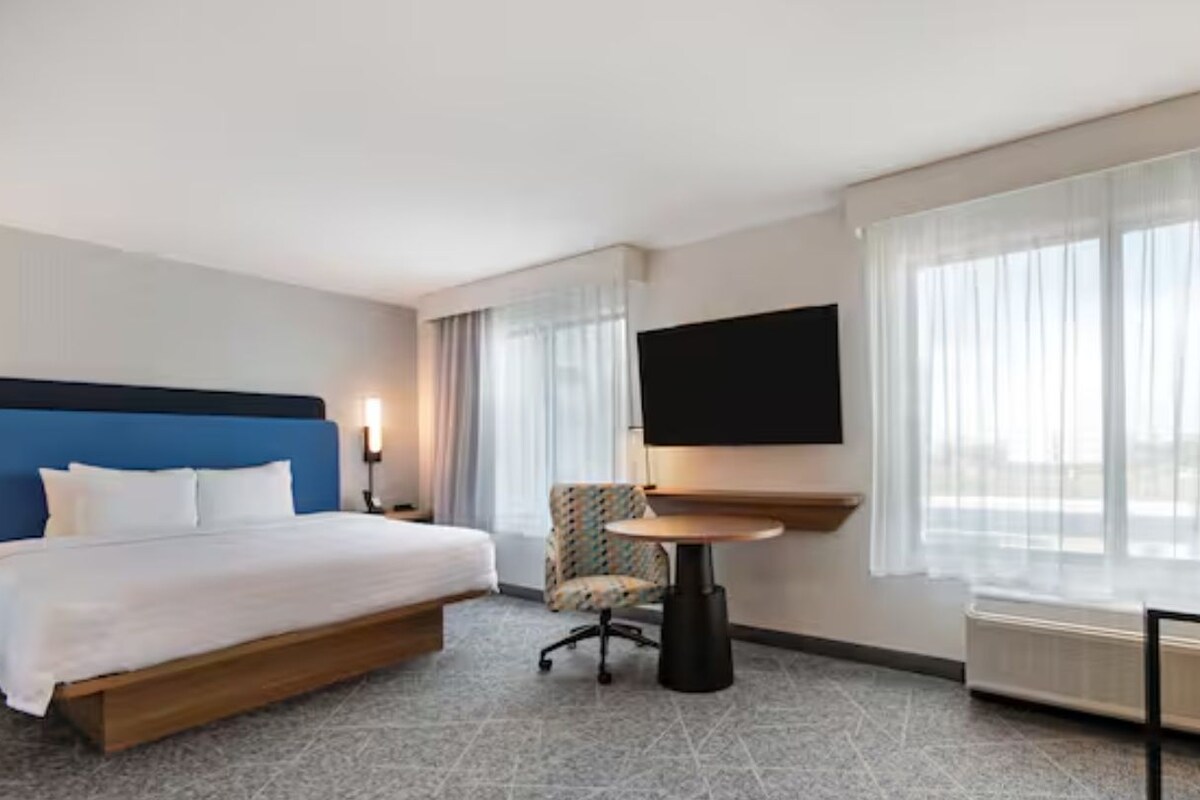 Habitación de hotel con cama, escritorio, silla, televisor y ventanas amplias.