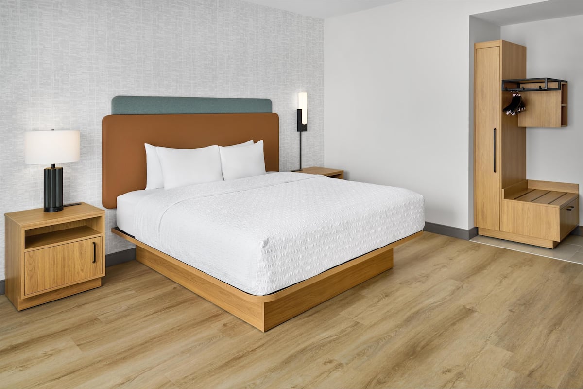 A modern hotel room with a wooden bed, a nightstand with a lamp, a wardrobe, and a textured wall.