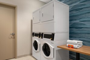 A laundry area with stacked washers and dryers, a wooden table with folded towels, and a door. - Hampton Inn By Hilton Lawrenceburg (Lawrenceburg)