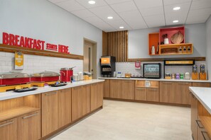 A breakfast area with a sign indicating breakfast starts here, a juice dispenser, and a coffee machine.