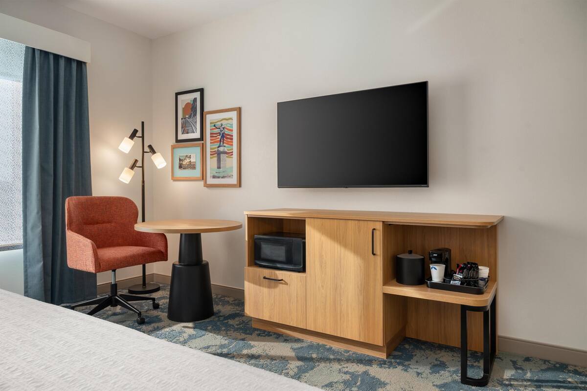 Habitación de hotel con cama, sillón rojo, una mesita, televisor, microondas, cafetera y cuadros en la pared.