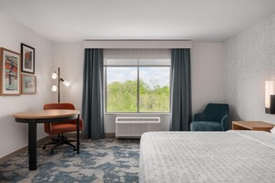 Habitación de hotel con cama, escritorio, silla, ventana con cortinas e imágenes enmarcadas en la pared.
