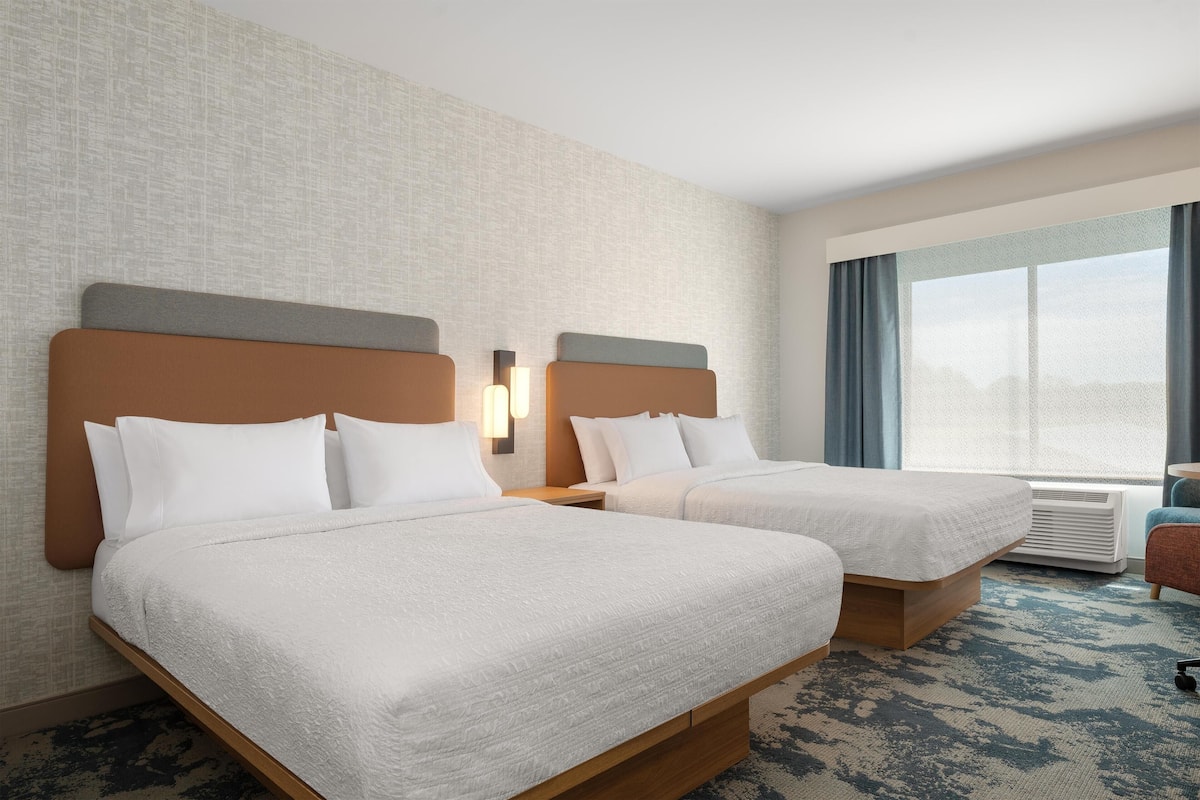 Una habitación de hotel con dos camas, un ventanal con cortinas, un aire acondicionado de pared y una alfombra estampada.