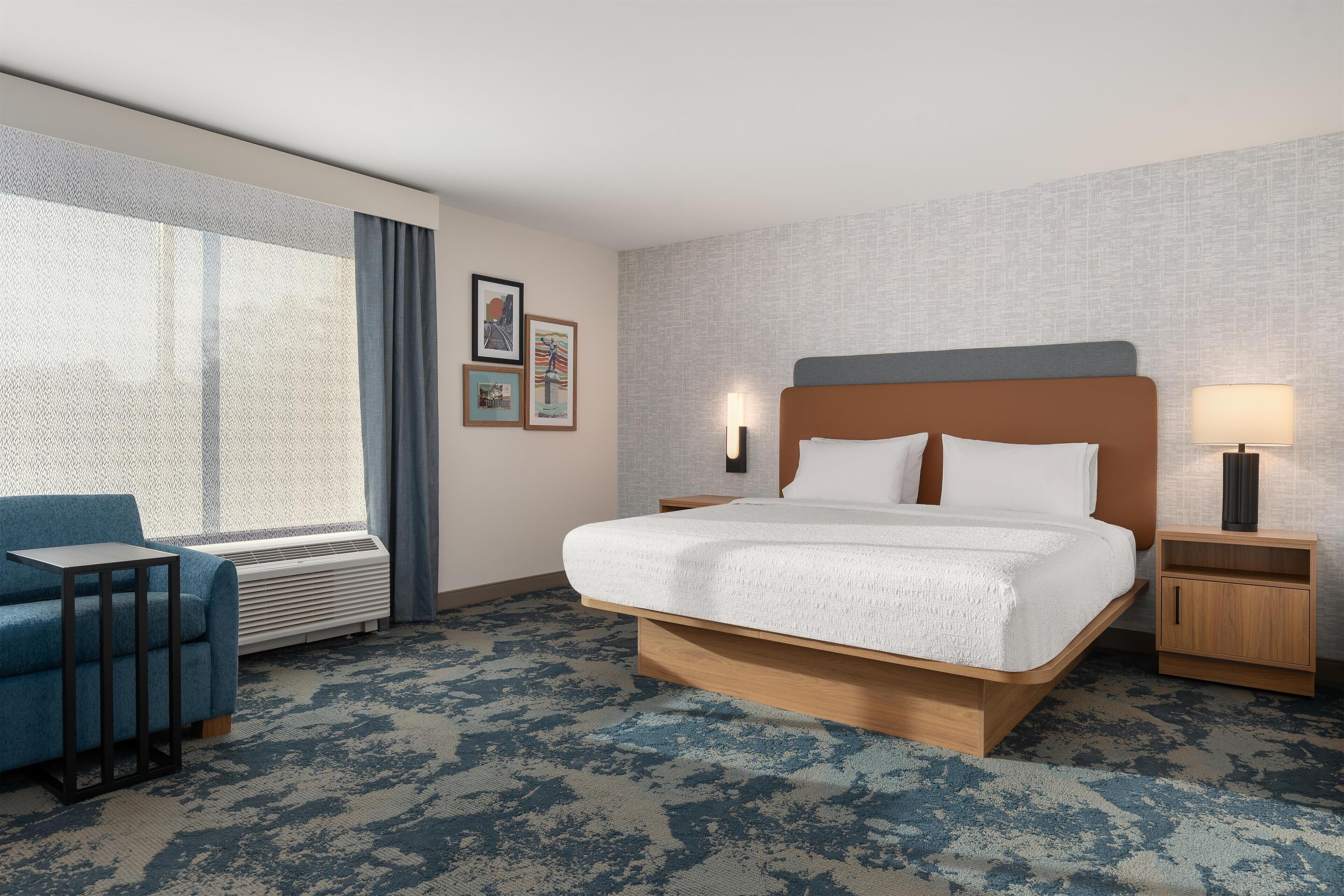 A hotel room with a large bed, a blue armchair, a bedside table with a lamp, and a window with curtains.
