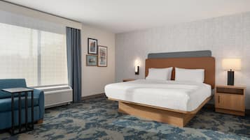 Ein Hotelzimmer mit einem großen Bett, einem blauen Sessel, einem Nachttisch mit Lampe und einem Fenster mit Vorhängen.