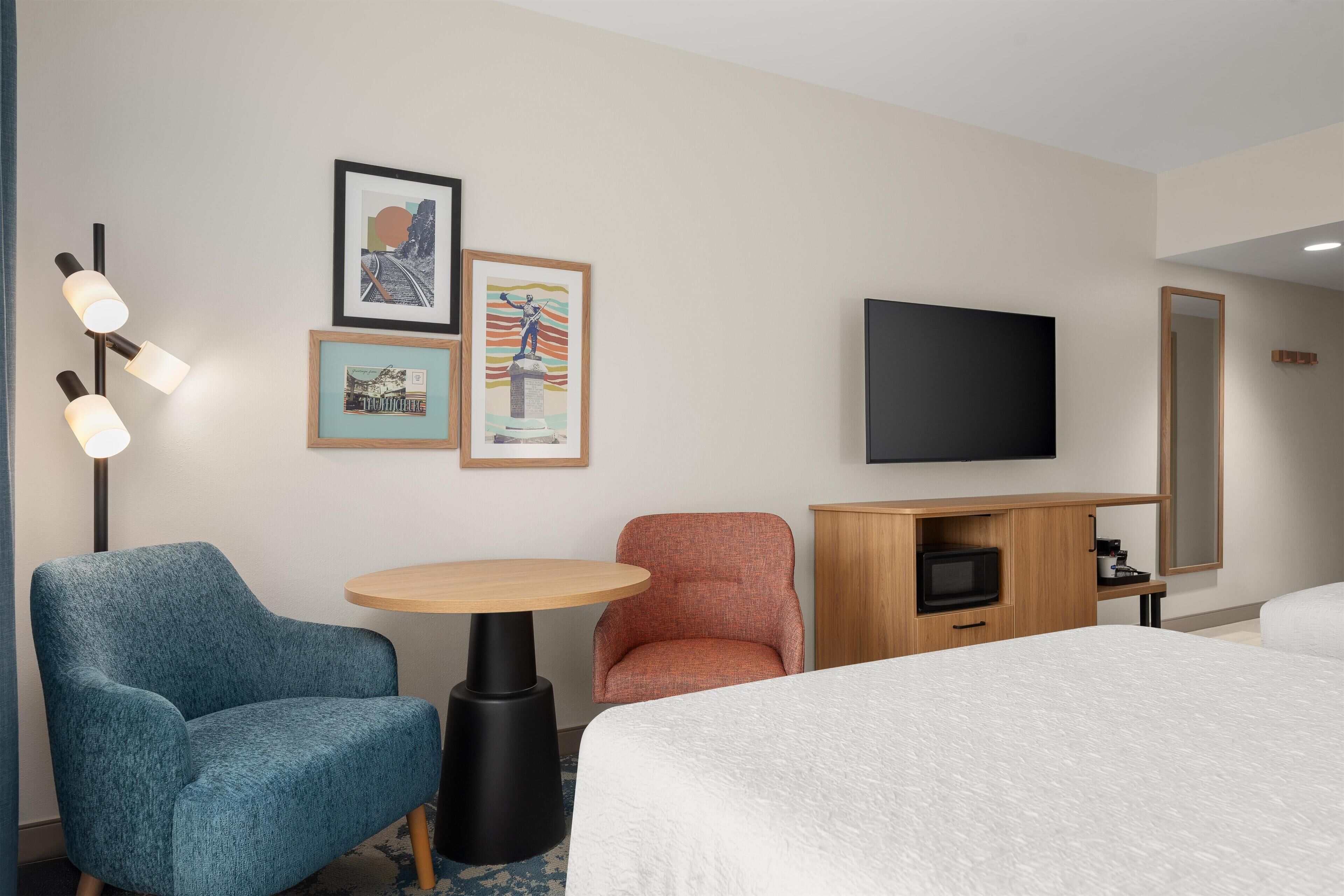 Ein Hotelzimmer mit einem Bett, einem blauen Sessel, einem Holztisch, einem Fernseher und gerahmten Kunstwerken an der Wand.