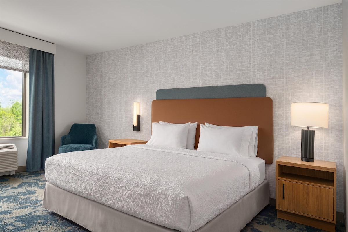 Habitación de hotel con una cama grande, una silla, una mesita de noche con lámpara y vistas a zonas verdes.