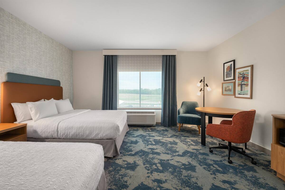 A hotel room with two beds, a desk, a chair, a lamp, and a window with curtains.