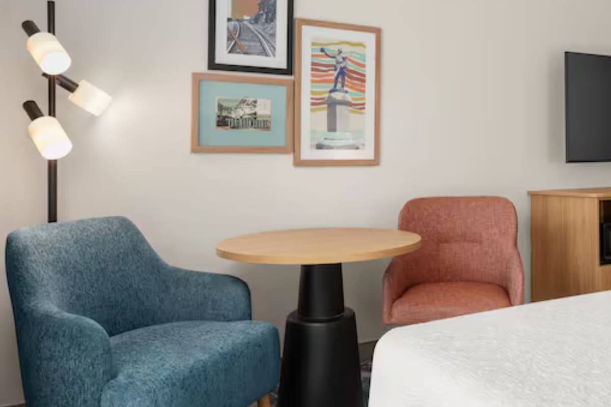 A hotel room with a bed, two chairs, a small table, a TV, and artwork on the wall.