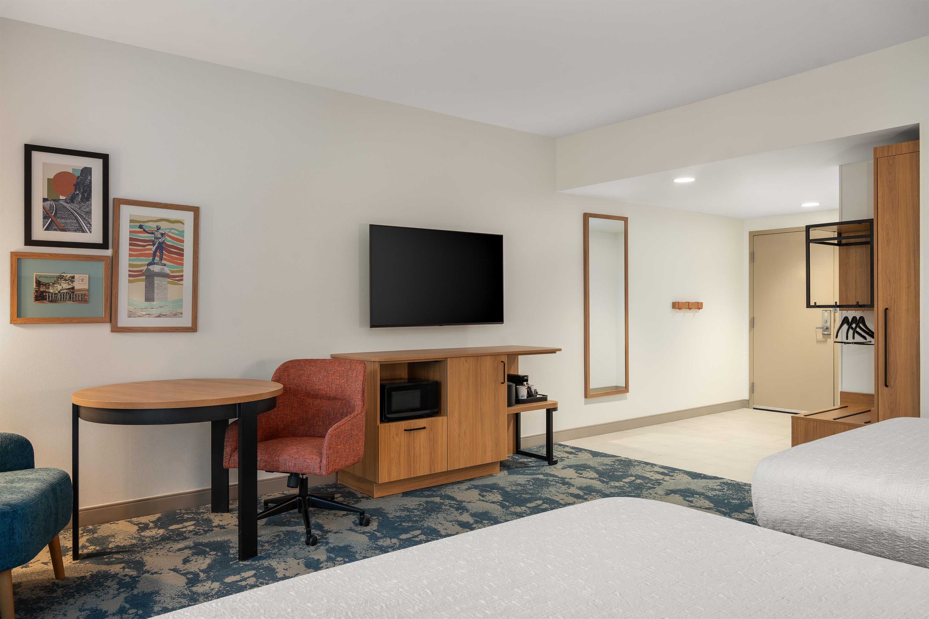 A hotel room with a bed, a desk with a chair, a television, a wardrobe, and framed pictures on the wall.