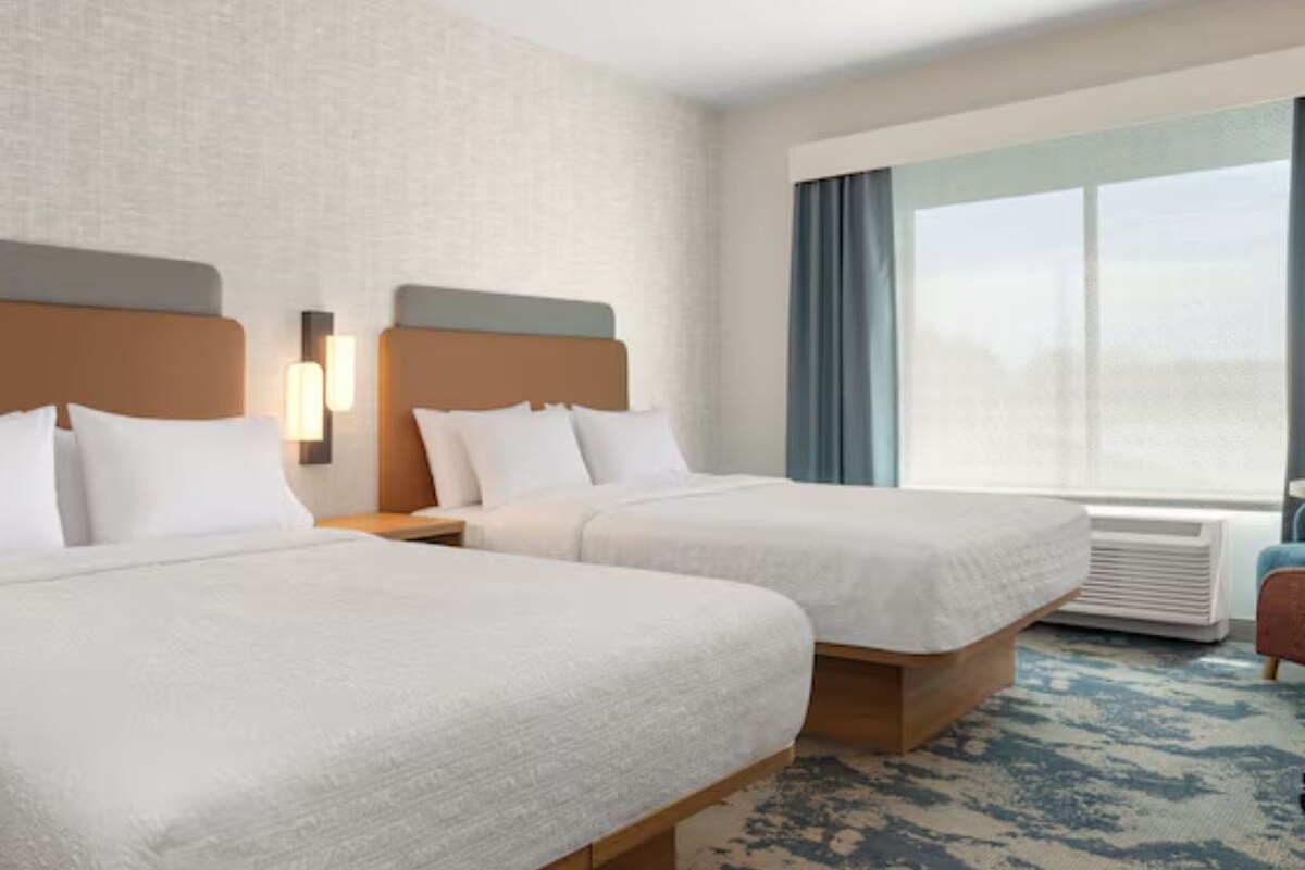 Ein Hotelzimmer mit zwei Betten, einem großen Fenster mit Vorhängen und einer wandmontierten Klimaanlage.