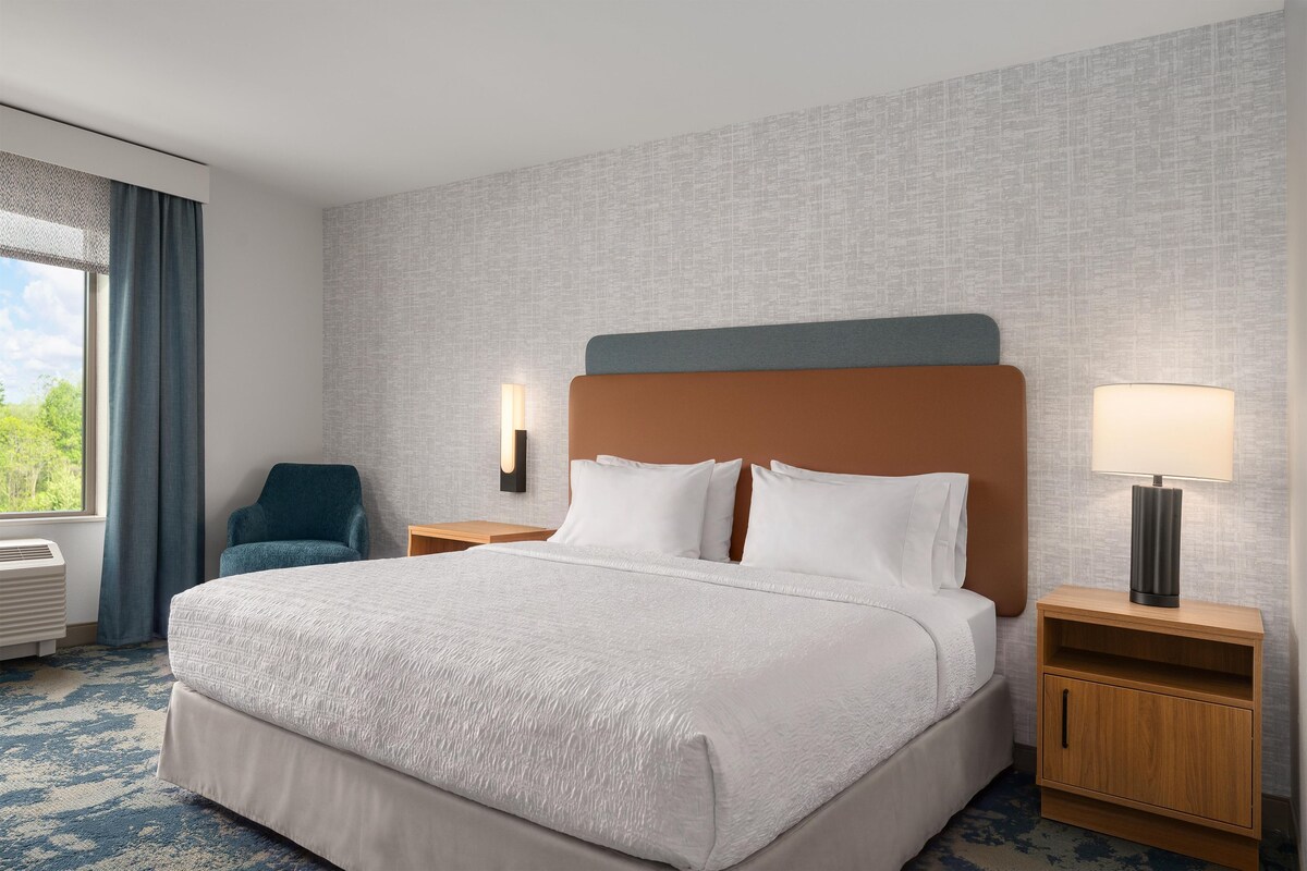 A hotel room with a large bed, a chair, a nightstand with a lamp, and a view of greenery outside.