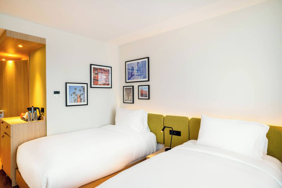 A hotel room with two beds, a wooden desk, and a nightstand. There are framed pictures on the wall.
