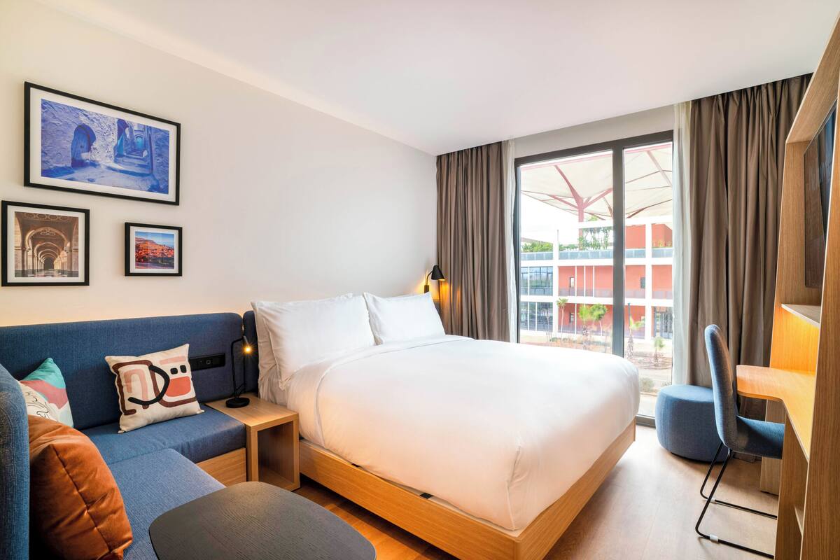 A modern hotel room with a bed, a desk, a chair, and a sofa. There are framed pictures on the wall and a window with curtains.