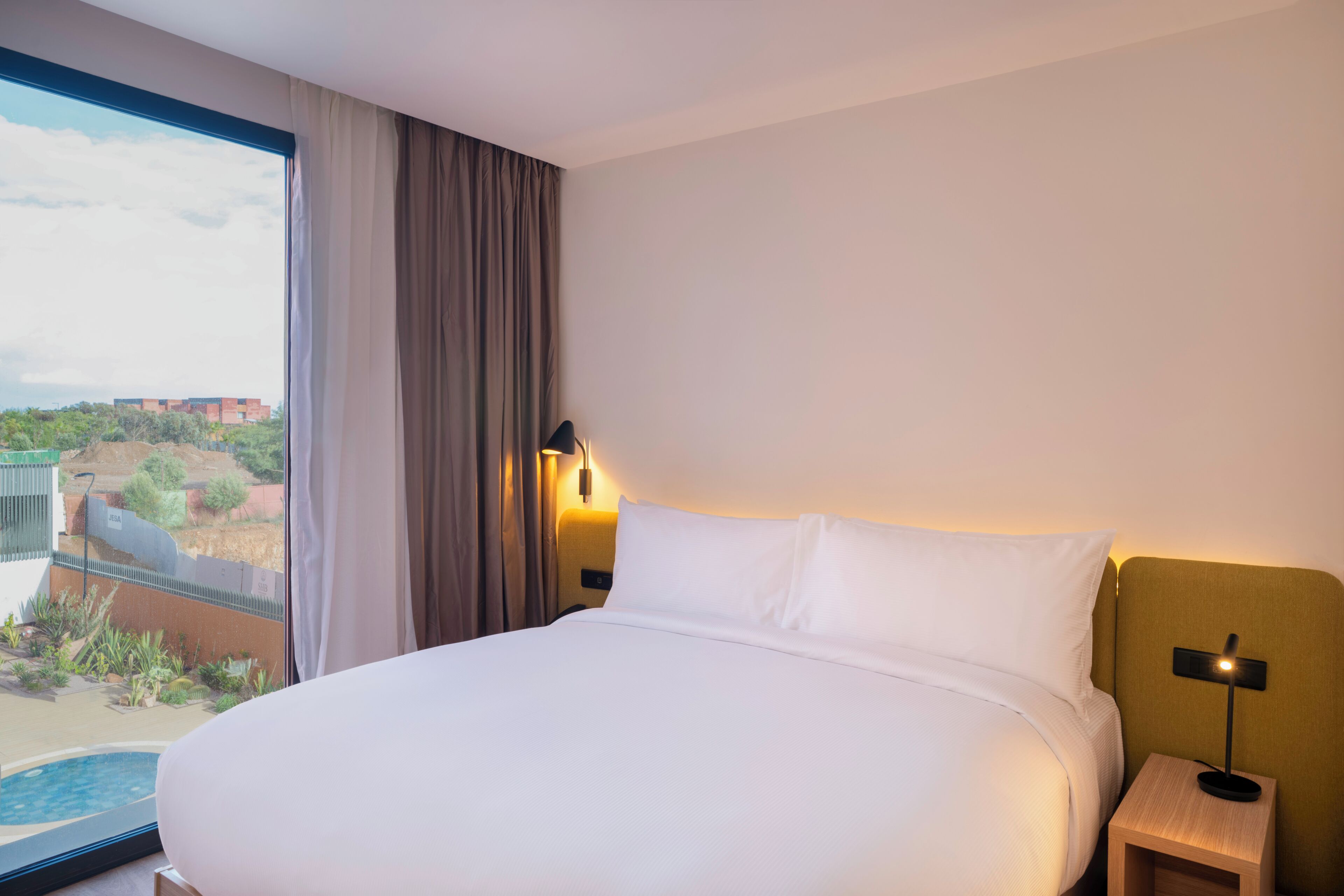 A hotel room with a large bed, a nightstand, a lamp, and a view of a swimming pool and buildings.