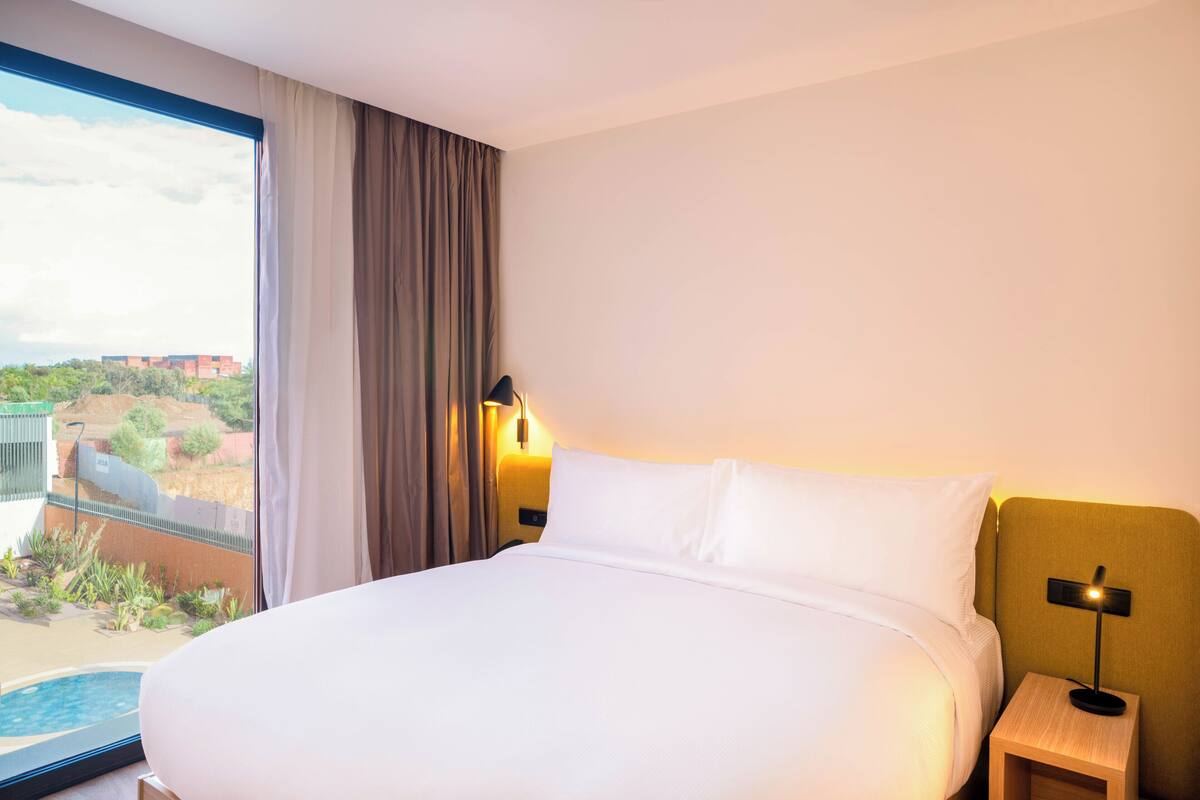 A hotel room with a large bed, a nightstand with a lamp, a view of a swimming pool, and a window with curtains.