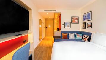 A hotel room with a large bed, a TV, a desk with a chair, and a sofa.