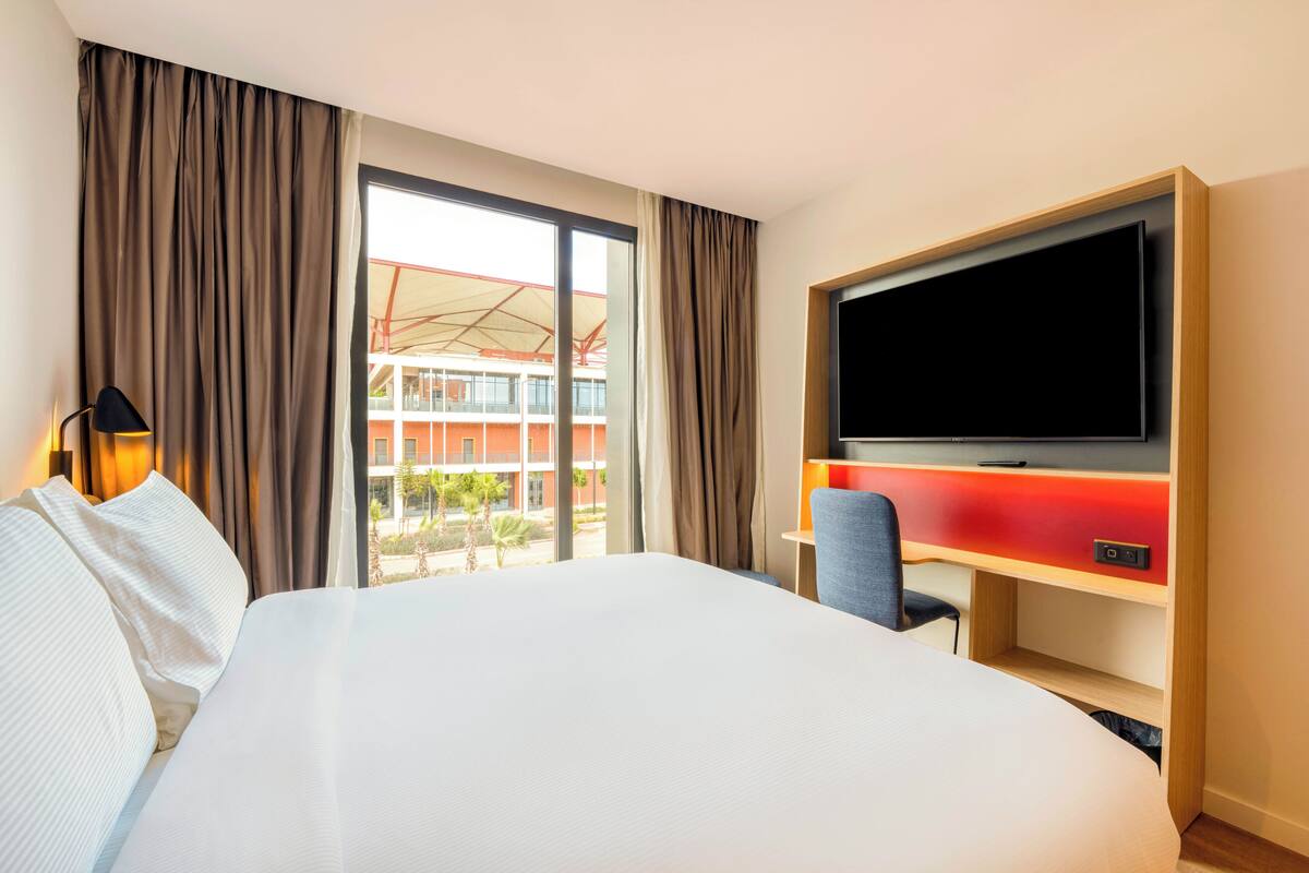 A hotel room with a large bed, a TV, a desk, and a view of a building outside.
