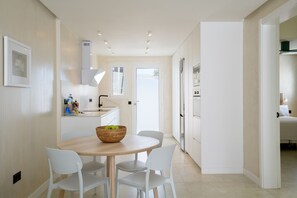 Een moderne keuken met witte kasten, een eethoek met een ronde tafel en witte stoelen, en een zichtbare koelkast.