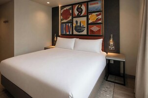 Habitación de hotel con una cama grande, cabecero decorado con arte, mesitas de noche y una lámpara fijada a la pared.