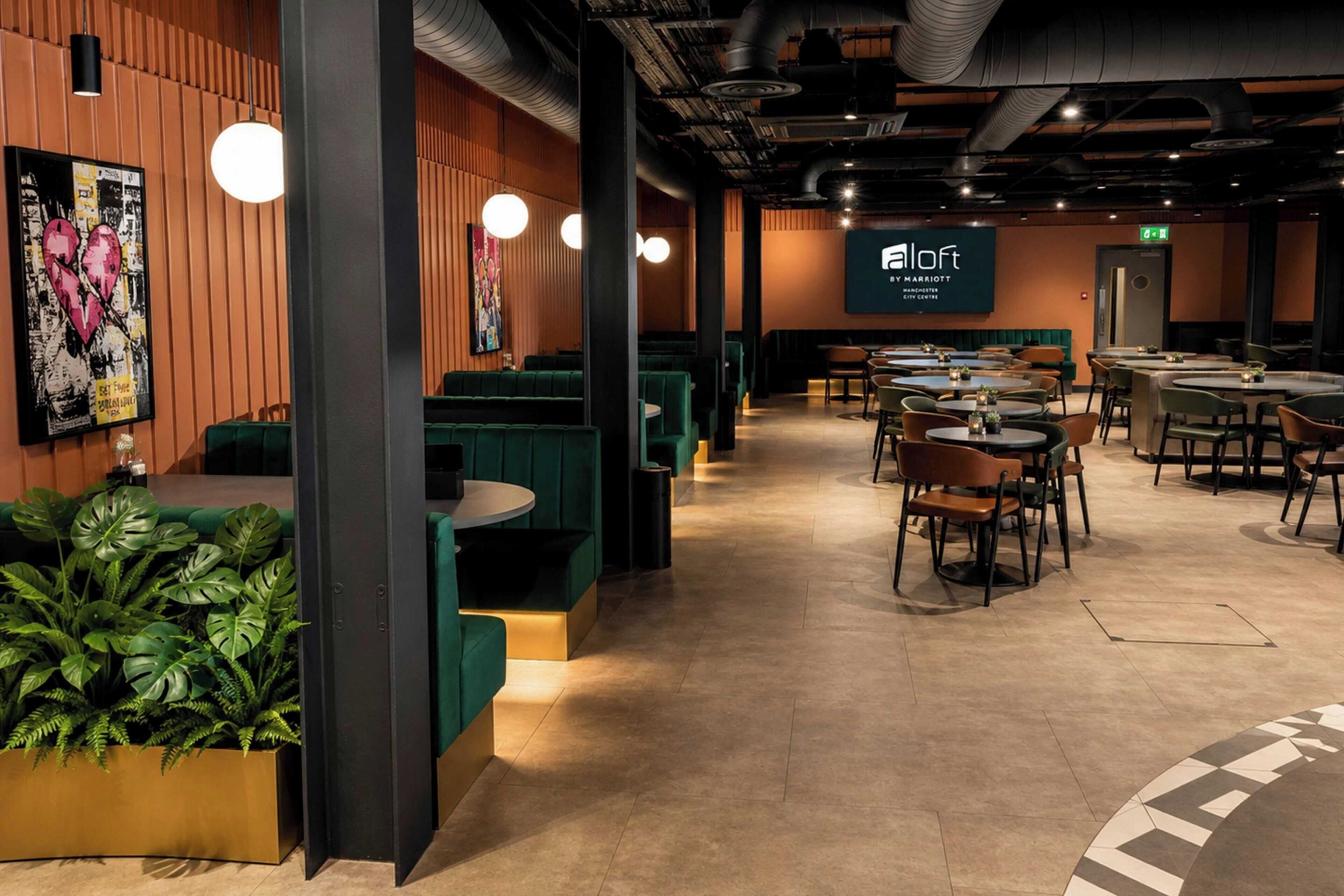Un restaurante moderno con asientos tipo banqueta verdes y mesas redondas, una pantalla grande que muestra el logotipo y una pared con una obra de arte con forma de corazón.