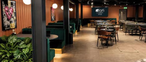 A modern restaurant with green booths and round tables, a large screen displaying the logo, and a wall with a heart-shaped artwork.